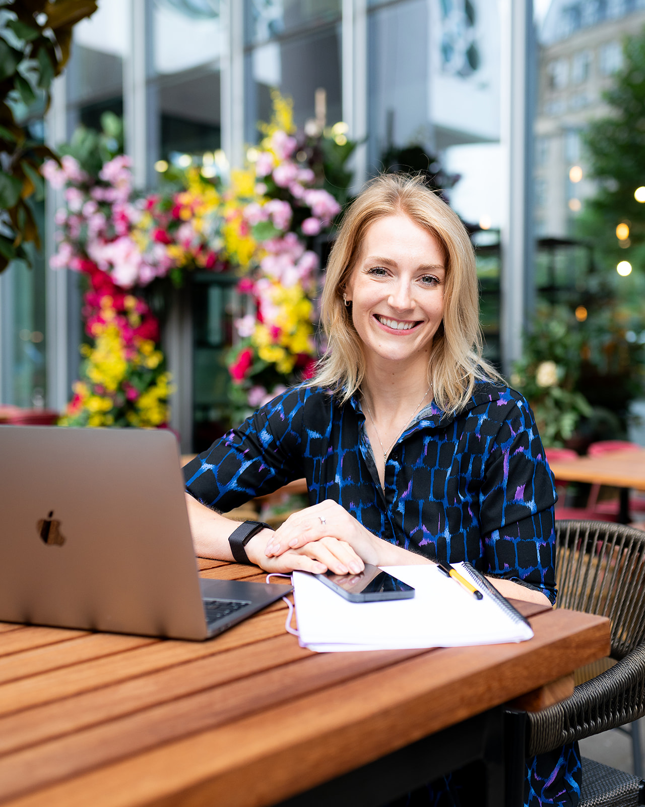 Kathy Brooke smiling at a table with her laptop and notebook, bright flowers behind her